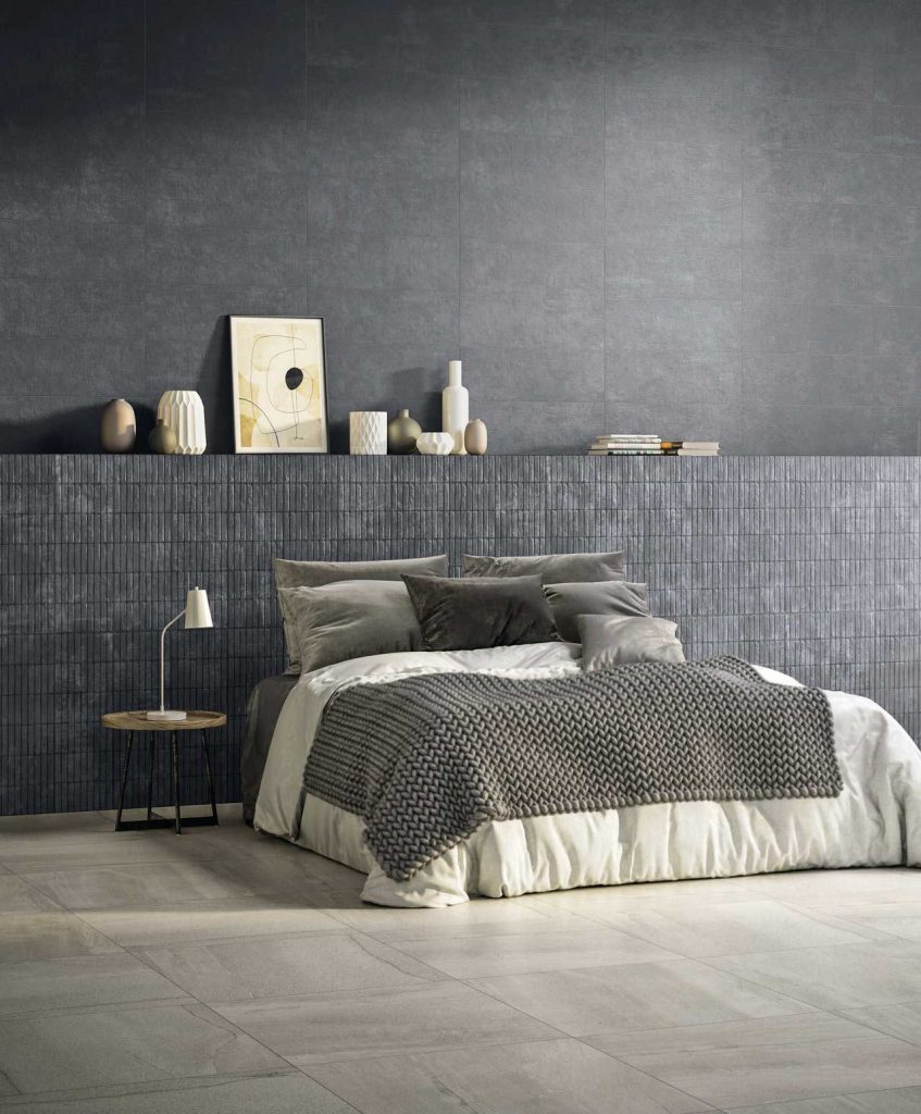 image showing active surfaces porcelain tiles being used in a bedroom on the floor and wall panelling, with a beige floor tile and grey wall tiles, with a grey double bed, side table and lamp, as well as several books and vases to decorate