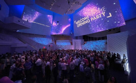 opening party in the new world center packed with attendees, screens above the hall display text saying welcome to the opening party