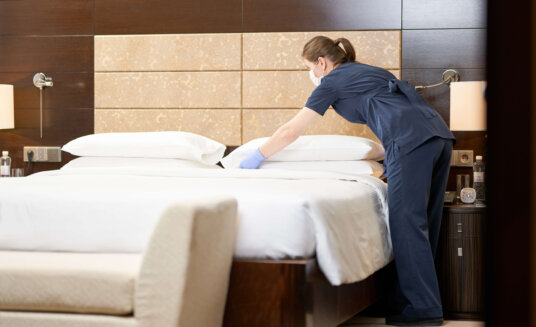 Full length photo of maid making bed and changing bedding in bedroom. Hotel service concept